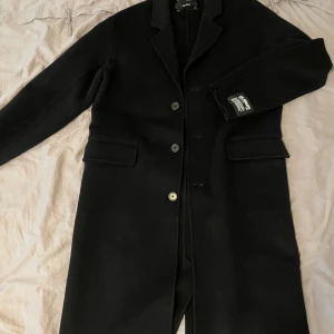 Svart rock från Axel Arigato - Snygg svart rock från Axel Arigato med klassisk krage, tre knappar framtill och två fickor med lock. Lång modell som ger en clean och stilren look. Perfekt för dig som vill ha en tidlös jacka med modern edge. Aldrig använd då den är för stor på mig, storlek L. Passar både kvinnor och Män😍