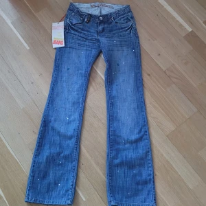 Low rise Musketeer jeans - Snygga blåa bootcut jeans från Musketeer med unika detaljer och stänkdesign i storlek 26. De är helt nya med lappar kvar! Om ni har några funderingar är det bara att fråga🩷 Tyvärr är de för små för mig så kan inte skicka bild med dem på. Midja: ca. 34. Innerbenslängd: ca 83 cm. Knappen som man stänger byxorna med är lite trasig, jag kan skicka bild om det behövs!