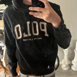 Svart hoodie från Polo Ralph Lauren - Svart hoodie från Polo Ralph Lauren med stor vit och beige POLO-logga på bröstet. Tröjan har huva med snörning och ribbade muddar vid ärmslut och nederkant. Mjuk insida i bomullsmix, perfekt för chill dagar. Klassisk och stilren design som alltid funkar. Passar 170-178, Nypris ca 2300