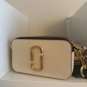 Marc Jacobs beige axelväska med guld - Snygg axelväska från Marc Jacobs i beige och svart med bred, randig axelrem och stora guldfärgade detaljer. Väskan har två fack med dragkedja och är tillverkad i skinn. Perfekt för dig som gillar stilrena accessoarer med statement-detaljer. Säljer på grund av ingen användning. Använt max 2 till 3 gånger (kan sänka priset vid snabb affär) 