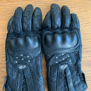 Svarta Held Lady motorcykelhandskar - Säljer ett par svarta Held Lady Equipment motorcykelhandskar i skinn med hårda knogskydd, dekorativa sömmar och diskreta mönster på fingrarna. Justerbara kardborreband vid handleden och förstärkta partier för extra skydd. Perfekt för dig som vill ha både stil och säkerhet på vägen.