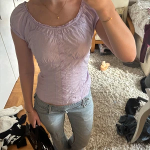 Ljuslila blus med knappar - Supersöt ljuslila blus med korta ärmar och rundad hals. Blusen har knappar framtill och fina rynkade detaljer vid halsen. Passformen är slim och materialet känns lätt och skönt mot huden, perfekt till jeans eller kjol för en chill look.