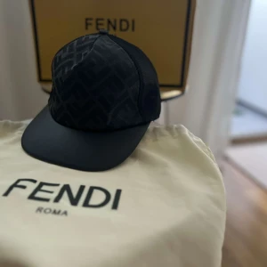Svart Fendi keps med FF-mönster - Snygg svart keps från Fendi med ikoniskt FF-mönster på framsidan och mesh-paneler på sidorna och bak. Justerbar rem med guldfärgad Fendi-logga baktill. Perfekt för dig som vill ha en lyxig och stilren accessoar.