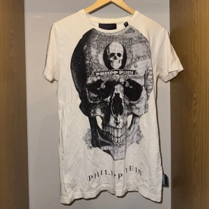 Vit t-shirt med döskalle från Philipp Plein - Snygg vit t-shirt från Philipp Plein med stor döskalleprint framtill och glittriga detaljer. Kortärmad modell i mjuk bomull. Logotypen är tryckt både fram och bak i nacken med en svart badge. Perfekt för dig som gillar edgy streetstyle.