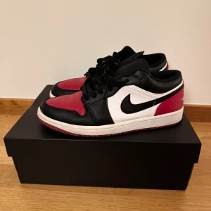 Nike Air Jordan 1 Low röd/svart/vit - Nike Air Jordan 1 Low sneakers med ikonisk färgblockning i rött, svart och vitt. Skorna har snörning, rund tå och platt sula. Ovandelen är tillverkad i skinn med broderad Air Jordan-logga på hälen och klassisk Swoosh på sidan. 