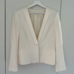 Beige kavaj från Filippa K, stl L - Elegant, ljus kavaj från Filippa K. Kavajen har en stilren design med en djup v-ringning och markerade axlar med axelvaddar. Perfekt för både vardag och fest. Finns nån liten fläck. Tight passform och lite liten i storlek.