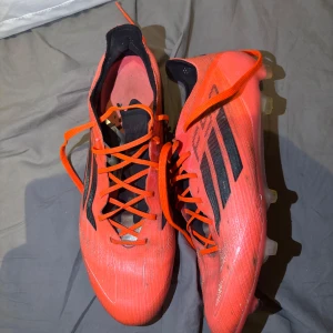Adidas X Speedflow röda fotbollsskor - Säljer ett par Adidas X Speedflow fotbollsskor i en riktigt snygg röd färg med svarta detaljer och orange snörning. Skorna har en spetsig tå och platt sula med dobbar för gräsplan. Ovandelen är tillverkad i syntetmaterial för lätt känsla och bra grepp.