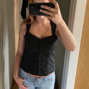 Svart prickig korsett-topp - Supersnygg svart korsett-topp med vita prickar och spetsdetalj vid bysten. Toppen är ärmlös och har en figurnära passform som framhäver siluetten. Perfekt att styla med jeans eller kjol för en cool look.