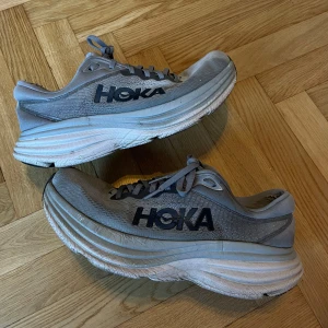Grå Hoka bondi 8 strl 43⅓ - Säljer ett par grå Hoka bondi 8 i storlek 43⅓, skicket är som på bilderna, köpta för 2199kr säljes nu för enbart 799kr. Sneakers med tjock, dämpad sula och ventilerande mesh-ovandel. Skorna har snörning och en sportig look med Hoka-logga i svart på sidan. Perfekta för dig som gillar att springa eller gå långt.