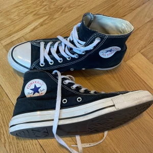 Svarta Converse Chuck Taylor High (storlek 41,5) - Klassiska svarta Converse Chuck Taylor All Star high tops med vit gummitå och sula i storlek 41,5 samt väldigt fint skick. Skorna har vit snörning, rund tå och tydliga vita sömmar. Märkets ikoniska logga på sidan och insidan är ljus. Perfekta för dig som gillar streetstyle och tidlös design.