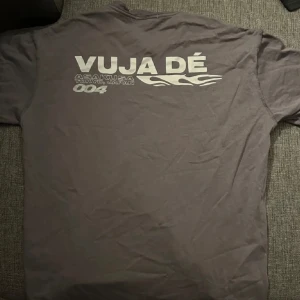 Vuja Dé t-shirt - Snygg mörkgrå t-shirt från Vuja Dé med coolt vitt tryck både fram och bak. Framsidan har en liten logga med flames och siffran 004, medan baksidan har ett större tryck med texten 'VUJA DÉ' och Asakusa Tokyo Japan. Perfekt för dig som gillar streetwear.