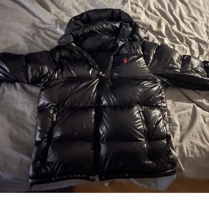 Svart pufferjacka Polo Ralph Lauren - Säljer en svart, glansig pufferjacka från Polo Ralph Lauren med huva och dragkedja. Jackan har två sidofickor med dragkedja och den klassiska röda Polo-loggan på bröstet. Perfekt för kalla dagar och riktigt snygg streetstil.