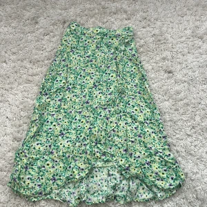 Blommig långkjol från Lindex Kids - Säljer en söt långkjol från Lindex Kids med blommigt mönster i grönt, gult, vitt och lila. Kjolen har volangkant nedtill och är gjord i ett mjukt, lätt polyestertyg som känns skönt mot huden. Perfekt för dig som gillar färgglada och lekfulla plagg!