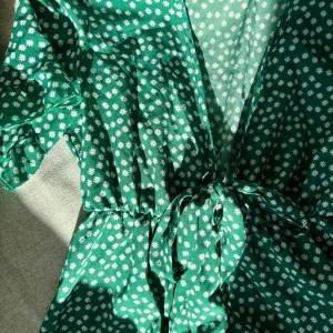 Grön blommig klönning med volanger - Säljer en grön klänning med vita blommor, korta ärmar och volangdetaljer. Klänningen har v-ringning och knyt vid bysten för en snygg passform. Perfekt för sommarens alla tillfällen, superfin färg och mönster som sticker ut. Använd fåtal gånger, fint skick! Vet tyvärr ej märket. Köpt via nätet. 