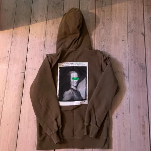 Brun hoodie från L'ADIEU VOLTAIRE - Säljer en brun hoodie från L'ADIEU VOLTAIRE med stor ficka framtill och tryck med konstmotiv och text på ryggen. Hoodien har huva och långa ärmar, samt diskret logga på bröstet. Perfekt för dig som gillar unik design och konstnärliga detaljer.