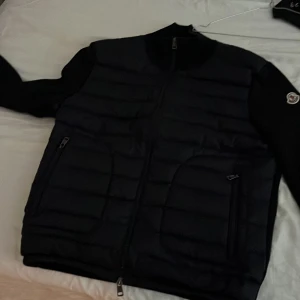 Svart pufferjacka från Moncler - Svart pufferjacka från Moncler med quiltad framsida och släta ärmar. Jackan har dragkedja framtill och två sidofickor med dragkedja. Klassisk Moncler-logga på ärmen och ribbad krage. Perfekt för dig som vill ha en stilren och exklusiv look.