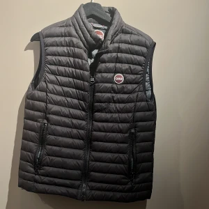 Svart dunväst från Colmar Storlek S - Snygg svart dunväst från Colmar med quiltad design och hög krage. Västen har två praktiska fickor med dragkedja och ett tydligt Colmar-märke på bröstet. Perfekt för lager-på-lager och chill stil under kyliga dagar.