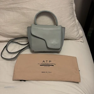 Ljusblå handväska från ATP Atelier - Säljer en ljusblå handväska från ATP Atelier med snygg asymmetrisk lockdesign och både handtag och axelrem. Väskan är tillverkad i skinn och har guldfärgade metalldetaljer. Kommer med dustbag. Perfekt storlek för det viktigaste. PRIS GÅR ATT DISKUTERA 