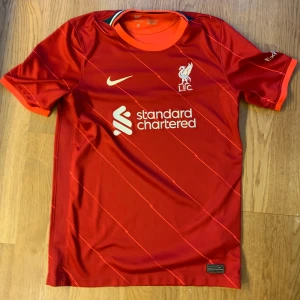 Liverpool FC röd matchtröja Nike XL Barn - Liverpool FCs officiella matchtröja i röd färg med subtila diagonala ränder. Tröjan har korta ärmar, vit logga och sponsortryck på bröstet. Tillverkad i lätt och ventilerande Dri-FIT polyester, perfekt för fotboll eller häng med polarna. Barnstorlek XL