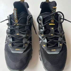 Nike React Element 55 sneakers svart/grå - Nike React Element 55 sneakers storlek 43 i svart och grått med gula detaljer. Ovandel i textil med rutmönster, grå och svarta syntetpaneler, och coola gula accenter på snörning och hälflik. Sulan är chunky och vågformad för extra streetkänsla. Perfekta för dig som gillar sportig och modern stil.