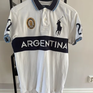 Vit Polo Ralph Lauren Argentina piké  - Vit kortärmad piké från Polo Ralph Lauren med blå detaljer, stor logga och texten ARGENTINA över bröstet. Snygg pikékrage med blå ränder, broderad emblem och nummer 2 på ärmarna samt 28 på ryggen. Tillverkad i bomull och har en sportig vibe. Limited edition piké
