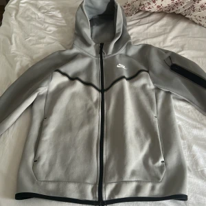 Grå hoodie från Nike med dragkedja - Snygg grå hoodie från Nike med svart dragkedja och huva. Tröjan har svarta detaljer längs bröstet och en ficka med dragkedja på ärmen. Perfekt för chill eller träning, med långärmad passform och mjukt material. Aldrig använd helt ny skick 