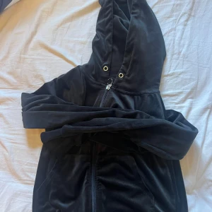 Svart velour hoodie från Gina Tricot - Mjuk och skön svart hoodie i velour från Gina Tricot. Tröjan har dragkedja framtill, huva med snörningshål och långa ärmar. Perfekt för en chill och avslappnad stil.