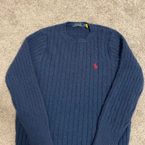 Marinblå kabelstickad tröja Polo Ralph Lauren - Kabelstickad marinblå tröja från Polo Ralph Lauren med klassisk röd broderad logga på bröstet. Tröjan har rund halsringning och långa ärmar, perfekt för lager-på-lager. Mjuk och skön ullblandning som håller dig varm under kyliga dagar.