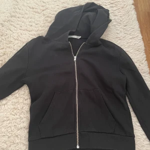 Svart hoodie med dragkedja från 157 - Svart hoodie från 157 i storlek S med hel dragkedja framtill och två stora fickor. Tröjan har huva och är tillverkad i mjuk bomullsmix, perfekt för chill dagar. Enkel och stilren design som funkar till det mesta.