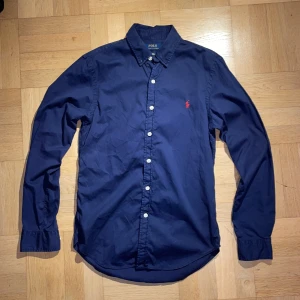 Mörkblå skjorta från Polo Ralph Lauren - Snygg mörkblå skjorta från Polo Ralph Lauren i bomull med klassisk krage och vita knappar. Slim fit-modell med röd broderad logga på bröstet. Perfekt för dig som gillar stilrena och tidlösa plagg. Svarar gärna på frågor och skickar inom 24h! 😊