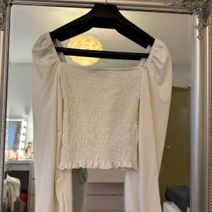 Vit smockad topp från Gina Tricot - Supersöt vit topp från Gina Tricot med smockad framsida och långa puffärmar. Fyrkantig ringning och lätt volangkant nedtill ger en trendig vibe. Perfekt att styla med jeans eller kjol för en fräsch look.