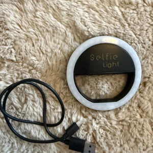 Selfie Light (ringlampa för mobil) - Selfie Light ringlampa i svart och vitt, perfekt för att få flawless ljus till selfies eller sminkvideos. Laddas med USB- c, vilket tillkommer. Har en klämma som går att sätta fast ovanpå telefonen.