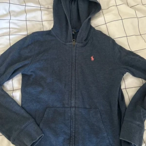 Mörkblå hoodie från Polo Ralph Lauren - (PASSAR S)Säljer en mörkblå hoodie från Polo Ralph Lauren i storlek XL. Tröjan har dragkedja, huva och en klassisk känguruficka framtill. På bröstet finns Polo-loggan i rosa. Perfekt för chill dagar och enkel att matcha med jeans eller joggers.