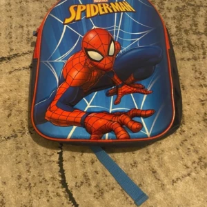 Blå Spiderman ryggsäck med röd dragkedja - Cool ryggsäck med Spiderman-tryck på framsidan, blå bakgrund och röda detaljer. Justerbara blå axelremmar och röd dragkedja. Perfekt för dig som gillar superhjältar och vill sticka ut med en färgstark väska. Materialet är slitstark polyester.