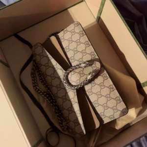 Beige Gucci Dionysus axelväska - Säljer en beige Gucci Dionysus axelväska med klassiskt GG-mönster och snygg silverfärgad kedja. Väskan har ett unikt spänne med två tigerhuvuden och är tillverkad i canvas med skinn-detaljer. Perfekt för dig som vill ha en ikonisk designerbag. Det är kristall detaljer på spännet vilket är en unik design, buda på pris ❤️ seriös säljare ser fram emot att höra från seriösa köpare, väskan är äkta har dock varken kvitto eller box kvar tyvär.. anledning till varför jag säljer är för den aldrig används 