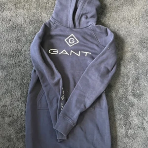 Blå klänning från GANT med logga - Snygg blå hoodie från GANT med stor vit logga framtill. Tröjan har huva, långa ärmar och en klassisk känguruficka. Perfekt för dig som gillar en avslappnad och sportig stil. Mjuk bomullskänsla och stilren design.