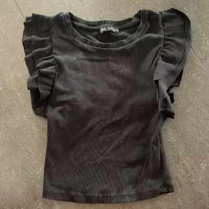 Svart topp med volang Zara - Svart ribbad topp från Zara med snygga volangärmar som ger en cool och trendig look. Toppen har rund halsringning och är figurnära i modellen. Perfekt att styla med jeans eller kjol för en stilren vibe.
