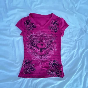 Rosa t-shirt med tryck och v-ringning - Cool rosa t-shirt från shein med svart tryck av vingar, rosor och hjärtan. T-shirten har v-ringad hals, korta ärmar och figurnära passform. Perfekt för dig som gillar edgy och detaljerade motiv.