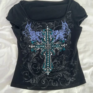 Svart topp med kors och strasstenar - Cool svart topp med korta ärmar och fyrkantig ringning. Framsidan har ett stort kors dekorerat med vita och turkosa strasstenar samt lila rosor och detaljerade mönster. Tight passform som ger en edgy vibe och sticker ut i mängden.