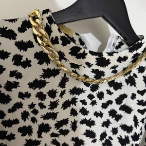 Svartvit blus med abstrakt mönster - Säljer en svartvit blus med coolt abstrakt mönster och lång ärm. Blusen har en hög krage och ett snyggt guldfärgat kedjehalsband som detalj. Materialet känns lätt och flowy, perfekt för dig som gillar att sticka ut med din stil.