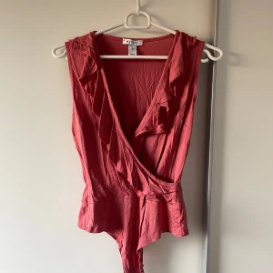 Röd omlott bodysuit från NLY One - Supersnygg röd bodysuit från NLY One med omlott-skärning och volangdetaljer längs urringningen. Ärmlös modell i mjukt bomullsmaterial, perfekt till jeans eller kjol för en trendig look.