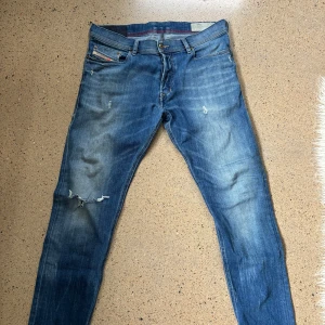 Diesel jeans herr - Snygga Diesel jeans i klassisk blå tvätt med slitningar och hål på vänster ben. Modellen har smal passform, fem fickor och dragkedja. Jeansen är gjorda i stretchig bomull för skön komfort och har diesel-logga på bakfickan.