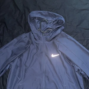 Nike Miller tracksuit thunder blue - Snygg marinblå tracksuit från Nike med huva och dragkedja framtill. Jackan har en clean look med reflekterande Nike-logga på bröstet och praktiska fickor. Perfekt för dig som gillar sportig stil och vill ha något lätt och smidigt.