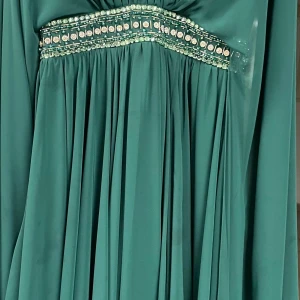 Grön långklänning med pärldetaljer - Elegant grön långklänning med glittrande pärldetaljer och stenar i midjan. Klänningen har lång ärm och ett vackert fallande tyg som ger en lyxig känsla. Perfekt för dig som vill sticka ut med en sofistikerad look.