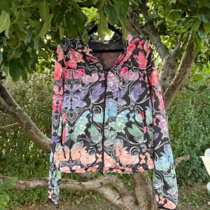 Färgglad vindjacka från Odd Molly - Snygg vindjacka från Odd Molly med färgstarkt fjärilsmönster i rosa, lila, blått, grönt och orange på svart botten. Jackan har dragkedja framtill, huva och långa ärmar. Perfekt för dig som vill sticka ut med en unik och lekfull stil. Storlek 2 som enligt deras storleksguide motsvarar en storlek M. Fint skick, men det finns tyvärr en anmärkning. Det är att dragkedjan på ena fickan är sönder. 