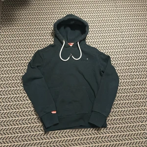 Svart hoodie från Superdry med huva - Svart Superdry hoodie med vit dragsko och broderad logga på bröstet. Klassisk känguruficka framtill och ribbade muddar. Mjuk insida och snygga detaljer som orange etikett på ärmen. Perfekt för chill dagar eller när du vill vara extra bekväm.
