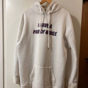Vit hoodie med vingar och text - Vit oversized hoodie med trycket 'I HAVE A PAIR OF WINGS' i glitch-stil på bröstet och stora rosa-svarta änglavingar på ryggen. Har huva med snörning och stor känguruficka framtill. Perfekt statement-plagg för dig som gillar unika prints.
