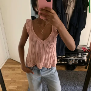 Ljusrosa stickad linnetopp - Supersöt ljusrosa stickad linnetopp med volangdetaljer vid axlarna och längst ner. Toppen har v-ringning och är ärmlös, perfekt för varma dagar. Den har ett luftigt, ribbat mönster som ger en chill och trendig vibe.