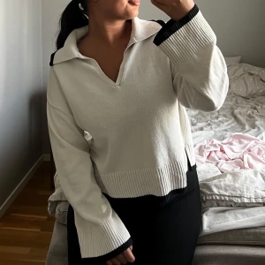 Vit stickad tröja med svarta detaljer - Vit stickad tröja med breda svarta ribbade kanter på ärmslut, krage och nederkant. Tröjan har en djup v-ringning och en avslappnad passform med långa ärmar. Perfekt för dig som gillar en stilren och trendig look.