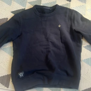 Mörkblå sweatshirt från Morris S - Snygg mörkblå sweatshirt från Morris med diskret logga broderad på bröstet och patch vid nederkanten. Klassisk rund hals och långa ärmar. Perfekt för dig som gillar stilrena och enkla plagg med en touch av premiumkänsla.